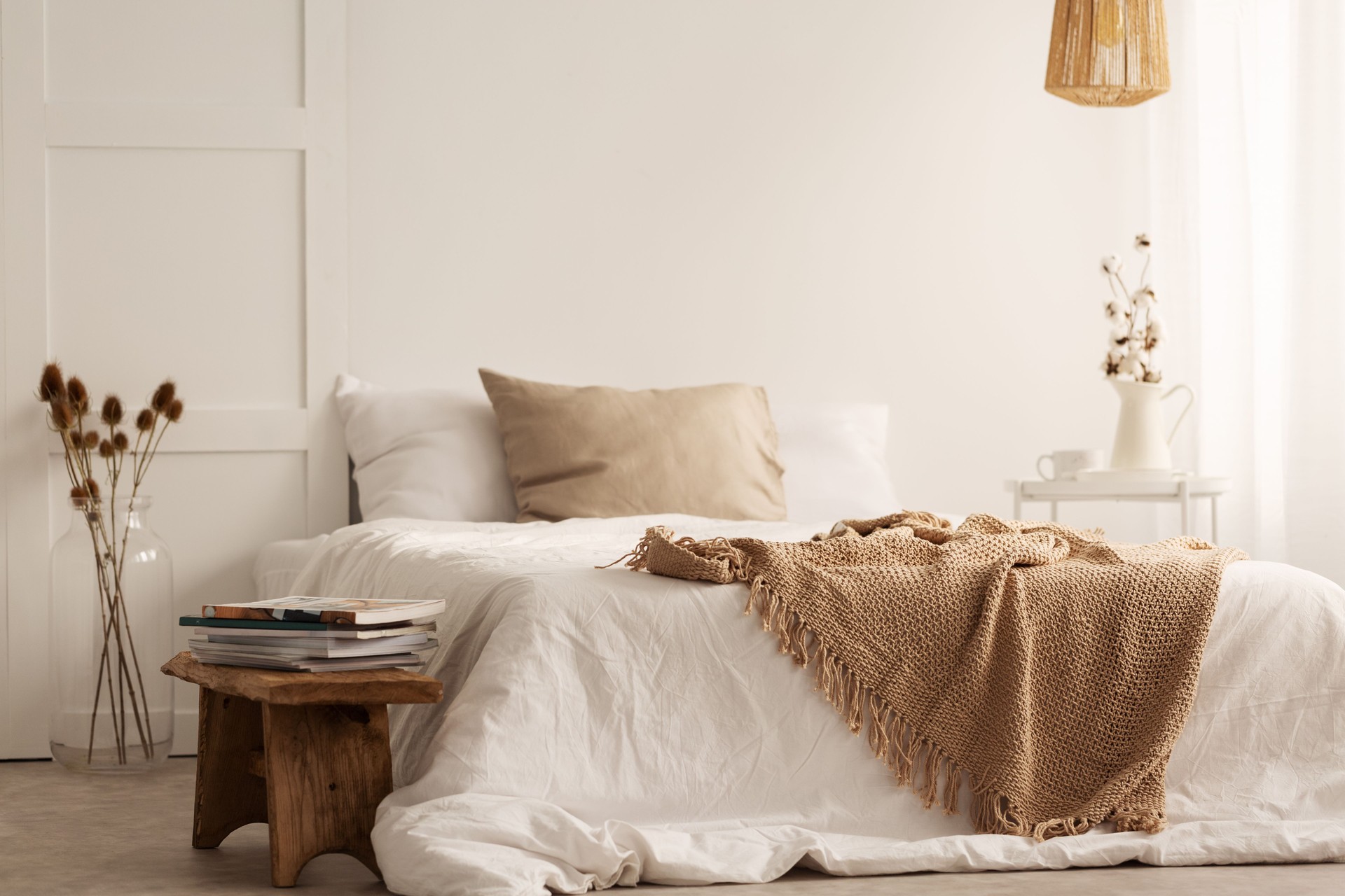 Blanket on white bed in natural bedroom interior with plants and wooden stool. Real photo